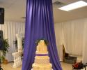 This gorgeous wedding cake table features beautiful purple and white accents to match the cake. The drapery behind the cake makes it stand out even more!